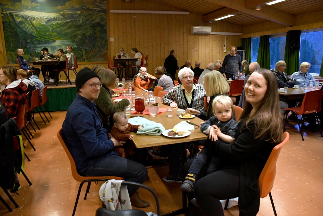 Far og mor og to små barn ved kafélangbord i framgrunnen, og diverse personar, unge og gamle i bakgrunnen