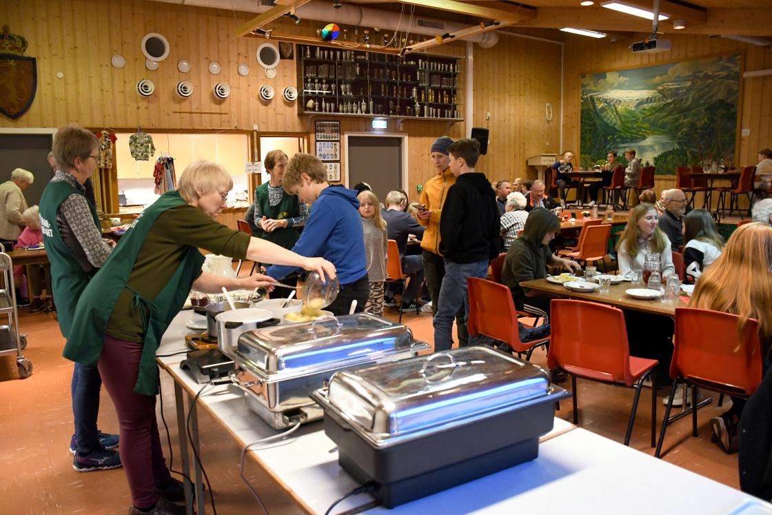Bygdekvinner i fine forkle etterfyller mat på serveringsbordet, med kafébord i bakgrunnen